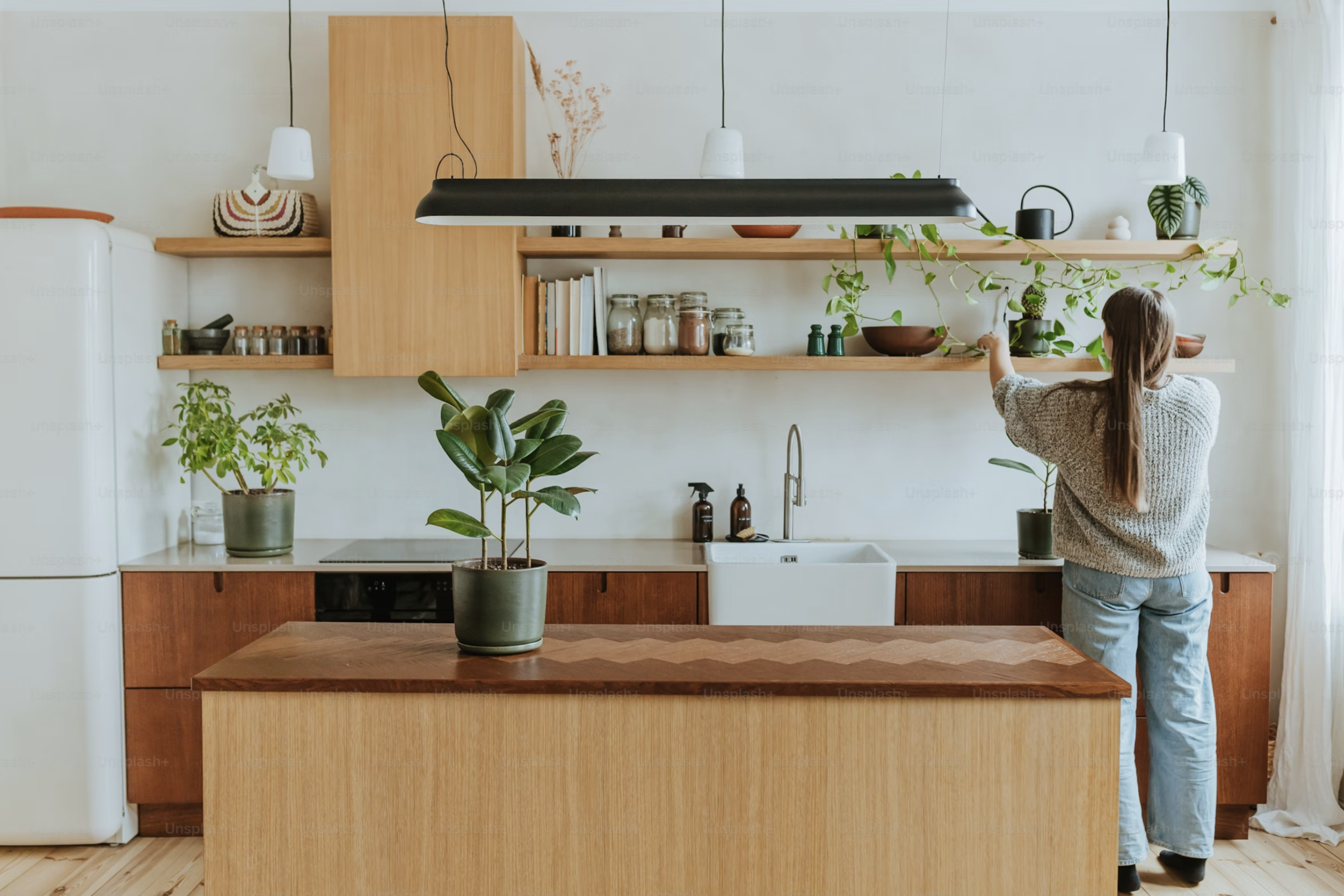 Person organizing items on wooden shelves in a bright, clean modern kitchen with white farmhouse sink, natural wood cabinetry, and green plants